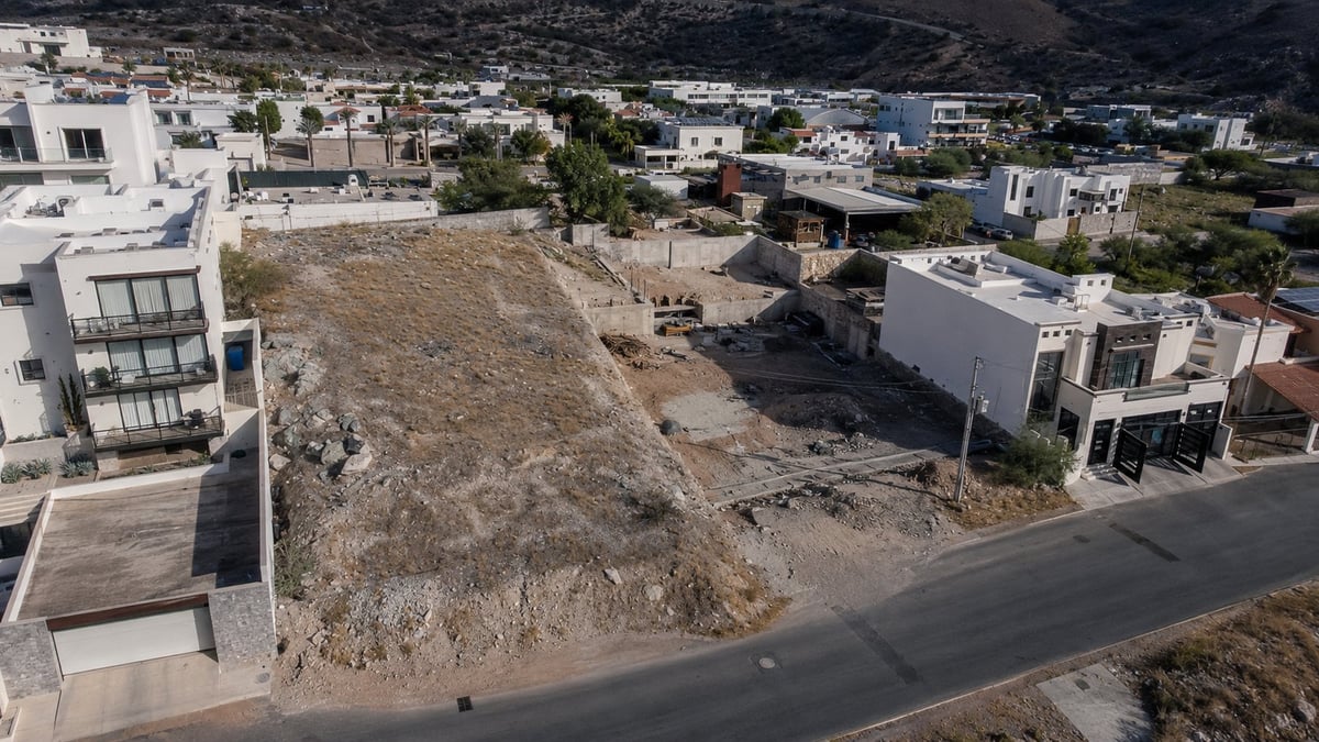 Aerial view of a construction site with cleared land surrounded by modern white residential buildings in a hillside neighborhood