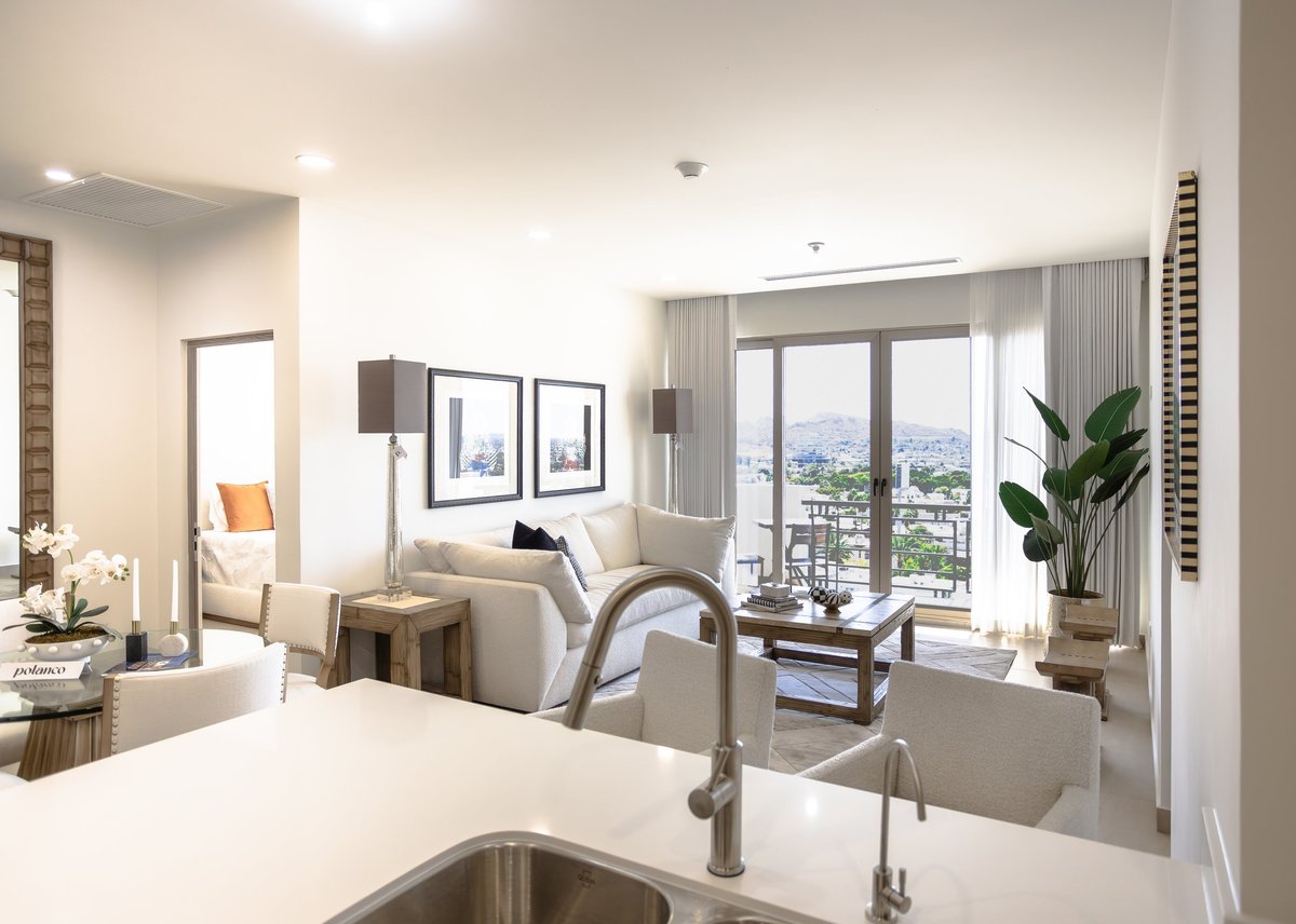 Modern kitchen and living room with stainless steel sink, white countertop, and floor-to-ceiling windows overlooking cityscape